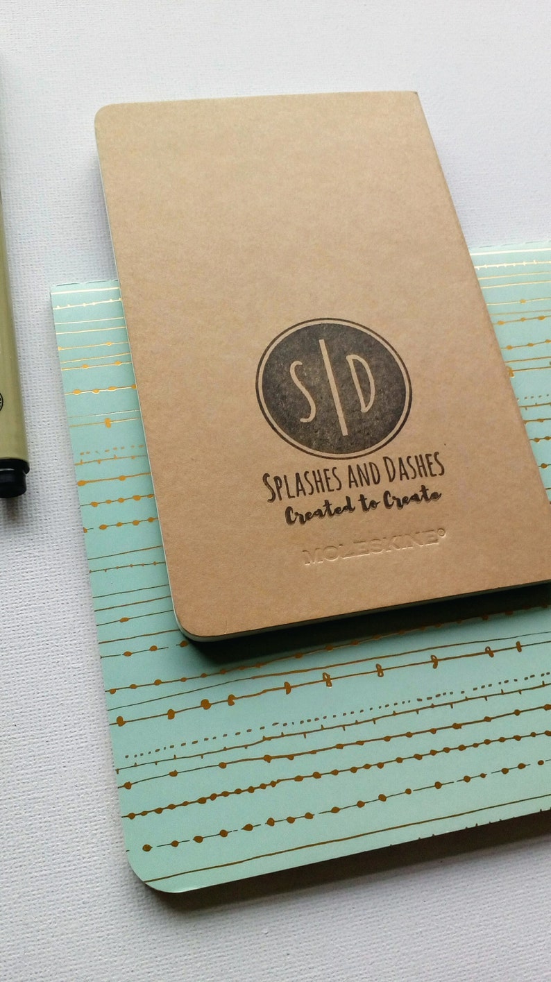 May include: A brown Moleskine notebook with a black circle logo that says "S|D" and the text "Splashes and Dashes Created to Create". The notebook is on top of a teal notebook with a gold pattern.