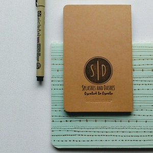May include: A brown notebook with the text "SPLASHES AND DASHES Created to Create" and a logo with the initials "SID" in a circle. The notebook is on top of a blue and white patterned notebook.