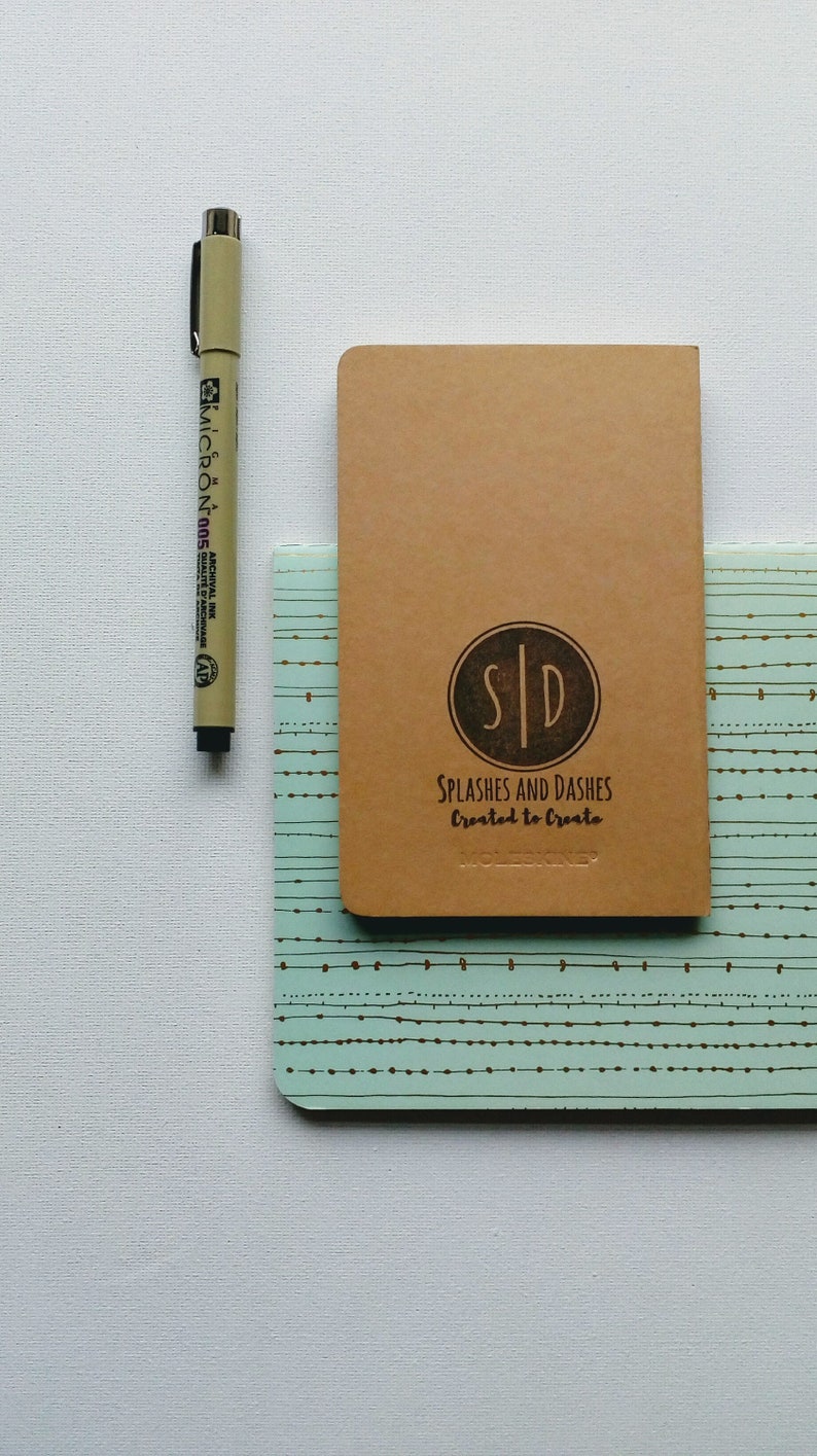 May include: A brown notebook with the text "SPLASHES AND DASHES Created to Create" and a logo with the initials "SID" in a circle. The notebook is on top of a blue and white patterned notebook.