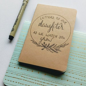 May include: A brown notebook with the text "Letters to our daughter as we watch you grow" written in black ink. The notebook is sitting on a light blue and gold patterned surface.