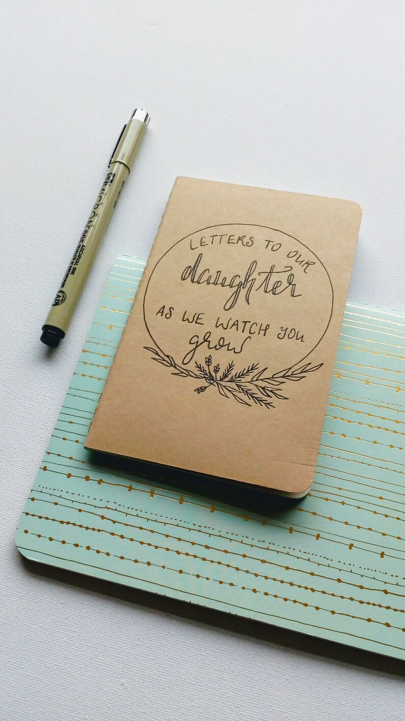 May include: A brown notebook with the text "Letters to our daughter as we watch you grow" written in black ink. The notebook is sitting on a light blue and gold patterned surface.