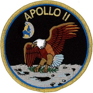 Może przedstawiać: Okrągła, haftowana naszywka z niebieskim i złotym obramowaniem. Naszywka przedstawia czarne tło z białym orłem lądującym na szarej powierzchni. Orzeł ma brązowe i pomarańczowe pióra. Księżyc jest widoczny w lewym górnym rogu. Tekst "APOLLO II" jest złotymi literami u góry naszywki.