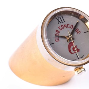 May include: A gold-toned desk clock with a white face and black Roman numeral hour markers. The clock face has the text "Club Conco de" in red lettering and a red crescent moon logo. The clock is on a gold-toned stand.