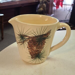 May include: A cream-colored ceramic pitcher with a brown pine cone and green pine needles design. The pitcher has a handle and a spout.