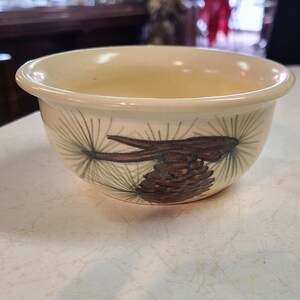 May include: A small, cream-colored ceramic bowl with a brown pine cone and green pine needles design.