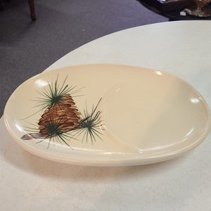 May include: A white oval ceramic serving dish with a divided section. The dish features a painted design of a brown pine cone with green pine needles.