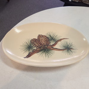 May include: An oval-shaped cream-colored ceramic platter featuring a central illustration of brown pinecones attached to a branch with green pine needles. The dish has a smooth, glossy finish and a slightly raised edge, showcasing a detailed botanical design.