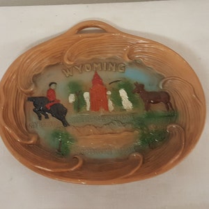 K&ouml;nnte beinhalten: Ein brauner Holz-Souvenirteller mit einem Reliefbild eines Cowboys, der ein Pferd reitet, einem roten Geb&auml;ude und einem Esel. Der Teller hat einen Griff und die Worte "WYOMING" sind in Wei&szlig; auf den Teller gedruckt.