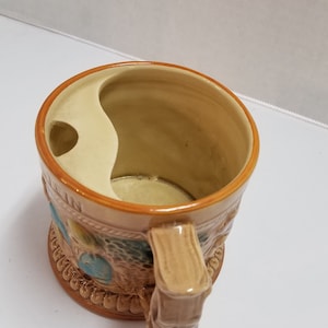 May include: A brown ceramic mug with a handle shaped like a tree trunk. The mug has a built-in shaving brush holder and features a decorative design of a forest scene.