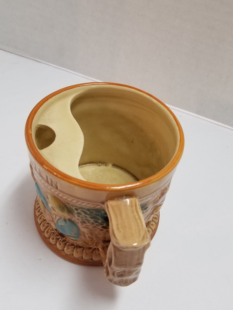 May include: A brown ceramic mug with a handle shaped like a tree trunk. The mug has a built-in shaving brush holder and features a decorative design of a forest scene.