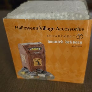 May include: A Halloween Village Accessories box featuring a miniature brown mailbox with a spooky design. The box is orange with a diamond pattern and the words "haunted delivery" are printed on the front.