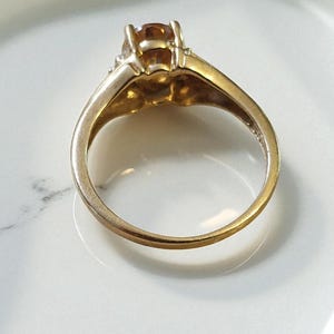 May include: A gold ring with a large, amber-colored gemstone. The ring is set on a white surface, with a white fabric in the background. The ring's band is a simple, classic design.