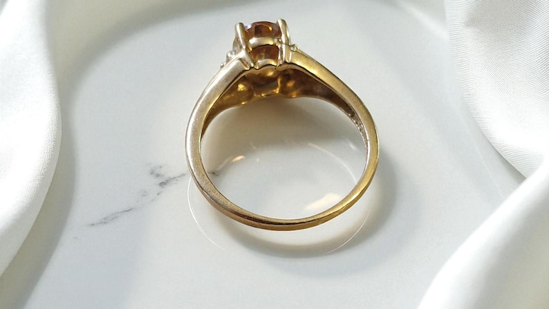 May include: A gold ring with a large, amber-colored gemstone. The ring is set on a white surface, with a white fabric in the background. The ring's band is a simple, classic design.