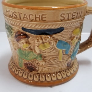 May include: A brown ceramic stein with a raised design of three men drinking beer. The stein has the words "Mustache Stein" on the front.