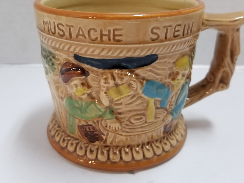 May include: A brown ceramic stein with a raised design of three men drinking beer. The stein has the words "Mustache Stein" on the front.