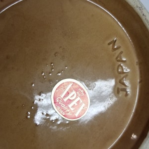 May include: Close-up of the bottom of a brown ceramic vase with a red and white "Apex Quality Japan" sticker. The vase is marked "Japan" in black lettering.