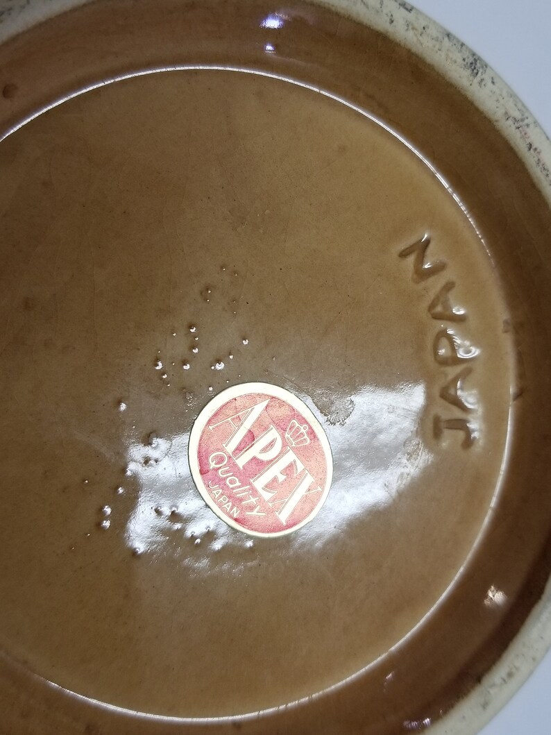 May include: Close-up of the bottom of a brown ceramic vase with a red and white "Apex Quality Japan" sticker. The vase is marked "Japan" in black lettering.