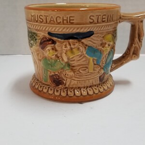May include: A brown ceramic stein with a tree-like handle. The stein features a scene of three men with mustaches drinking beer. The stein is decorated with a brown and white pattern and the words "MUSTACHE STEIN" are written on the front.