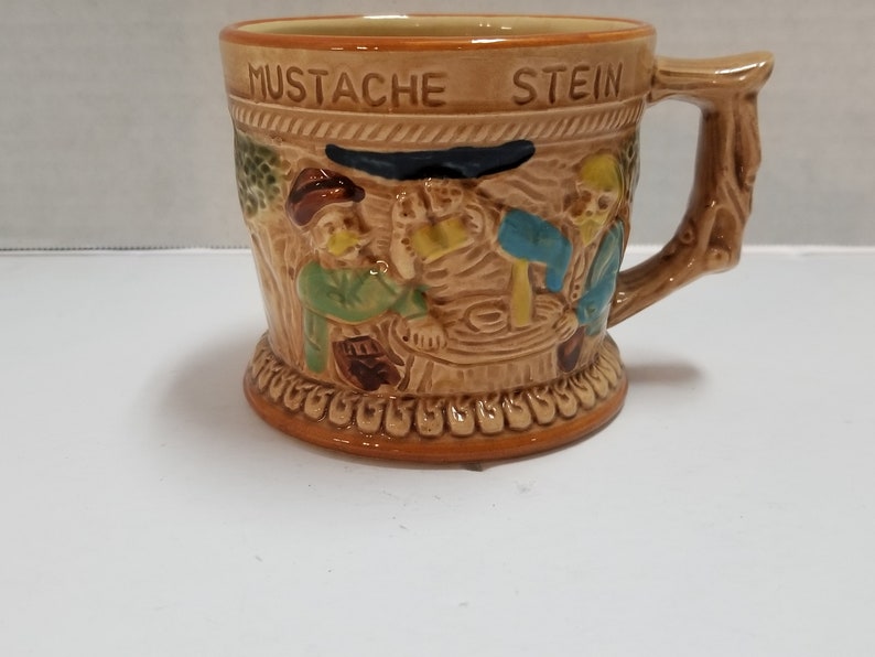 May include: A brown ceramic stein with a tree-like handle. The stein features a scene of three men with mustaches drinking beer. The stein is decorated with a brown and white pattern and the words "MUSTACHE STEIN" are written on the front.