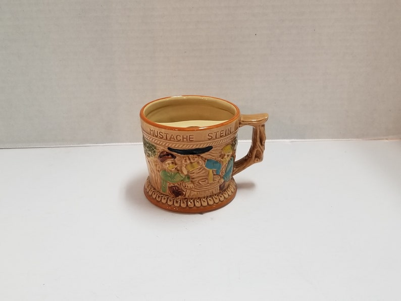 May include: A brown ceramic mustache stein with a handle shaped like a tree branch. The stein features a scene of three men with mustaches, one of whom is holding a beer mug. The stein is inscribed with the words "Mustache Stein."