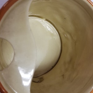 May include: A white ceramic mug with a brown rim and a built-in handle on the side. The mug has a small hole on the side.