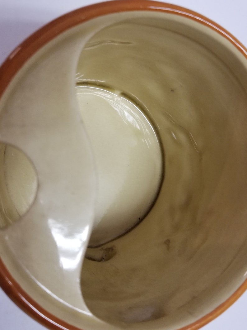 May include: A white ceramic mug with a brown rim and a built-in handle on the side. The mug has a small hole on the side.