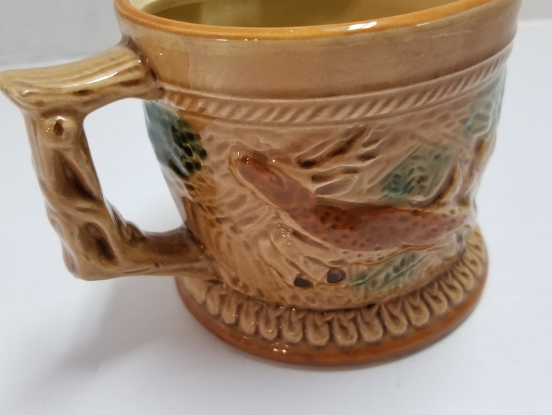 May include: A brown ceramic mug with a tree branch handle and a deer design. The mug has a brown glaze with green accents.