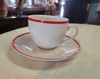 May include: A white ceramic coffee cup with a red band around the rim, sitting on a matching saucer with a red band around the edge.