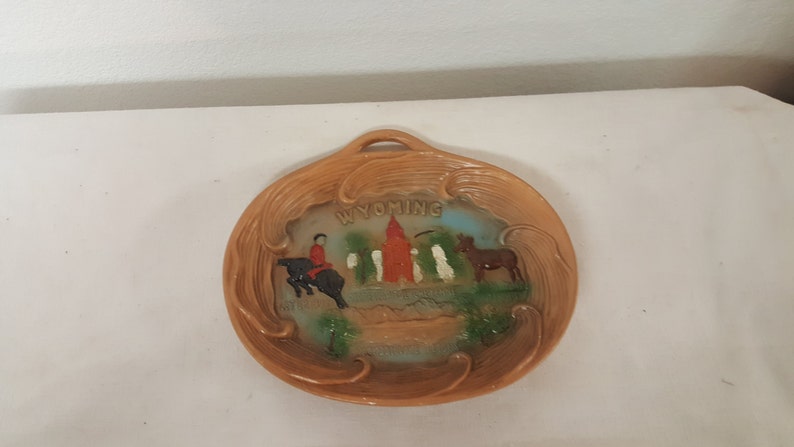 K&ouml;nnte beinhalten: Ein brauner Holz-Souvenirteller mit einem Reliefbild eines Cowboys, der ein Pferd reitet, einem roten Geb&auml;ude und einem Esel. Der Teller hat einen Griff und die Worte "WYOMING" sind in Wei&szlig; auf den Teller gedruckt.