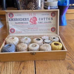 May include: A rectangular wooden box containing twelve assorted spools of embroidery cotton in various neutral and muted colors. The interior lid features a printed label with the text H.B. Embroidery Cotton, Guaranteed Fast Colors.