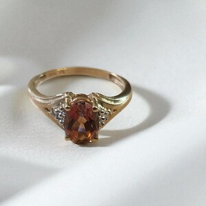 May include: A gold ring with an oval, amber-colored gemstone as the centerpiece. The ring has a classic design with small accent stones on either side of the main stone, set against a white, textured background.