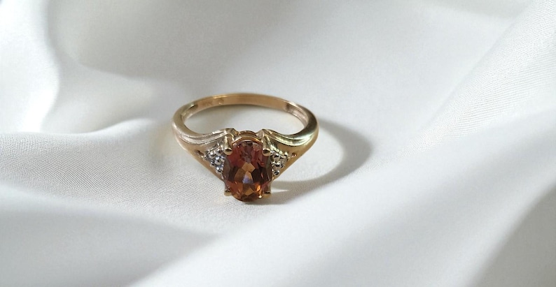 May include: A gold ring with an oval, amber-colored gemstone as the centerpiece. The ring has a classic design with small accent stones on either side of the main stone, set against a white, textured background.