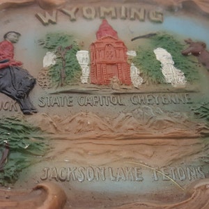 K&ouml;nnte beinhalten: Ein brauner und beigefarbener Aschenbecher aus Keramik mit einem erhabenen Design, das einen Cowboy auf einem Pferd, ein Staatskapitolgeb&auml;ude und eine Bergkette darstellt. Der Text "WYOMING", "STATE CAPITOL CHEYENNE", "LETTER BUCK", "JACKSON LAKE REGION" und "ANTELOPE" ist auf dem Aschenbecher aufgedruckt.