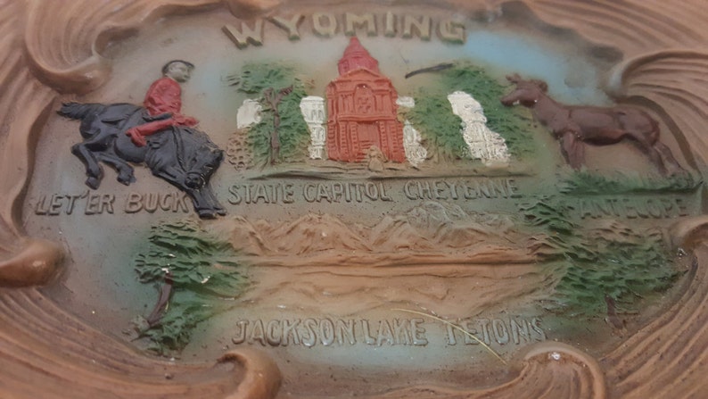 K&ouml;nnte beinhalten: Ein brauner und beigefarbener Aschenbecher aus Keramik mit einem erhabenen Design, das einen Cowboy auf einem Pferd, ein Staatskapitolgeb&auml;ude und eine Bergkette darstellt. Der Text "WYOMING", "STATE CAPITOL CHEYENNE", "LETTER BUCK", "JACKSON LAKE REGION" und "ANTELOPE" ist auf dem Aschenbecher aufgedruckt.