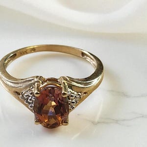May include: A gold ring with a large, oval, orange gemstone as the centerpiece. The ring has a classic design with small accent stones on either side of the main stone. The ring is set against a white marble surface.