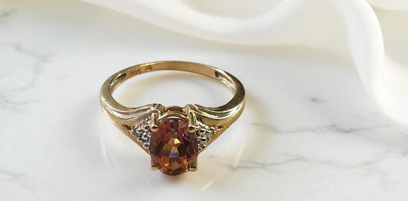 May include: A gold ring with a large, oval, orange gemstone as the centerpiece. The ring has a classic design with small accent stones on either side of the main stone. The ring is set against a white marble surface.