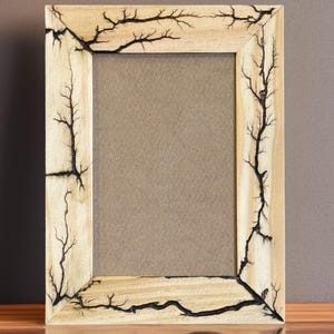Op de afbeelding: Een rechthoekige houten fotolijst met een natuurlijke houten afwerking. Het frame is versierd met zwarte, bliksemachtige patronen. Het frame staat op een reflecterend oppervlak.