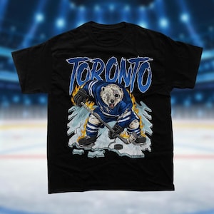 Peut inclure: T-shirt noir avec un graphique d'un ours polaire jouant au hockey, portant un maillot bleu et blanc. Le graphique comprend des flammes et le mot "TORONTO" dans une police de caractères stylisée.