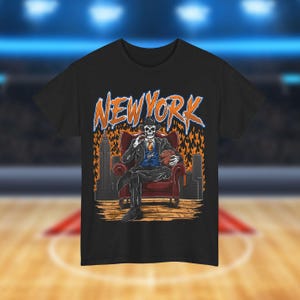 May include: Black t-shirt with a graphic of a skeleton wearing a suit and holding a basketball. The skeleton is sitting in a chair with flames behind it. The text "NEW YORK" is above the skeleton.