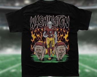 May include: Black t-shirt with a graphic of a skeleton football player in a red and gold uniform, running with a football. The player is surrounded by flames and two pigs wearing helmets are running behind him. The text "WASHINGTON" is printed above the player in a stylized font.