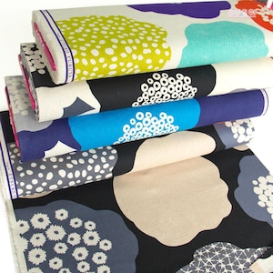 May include: Several rolls of patterned fabric in a stack. The designs include large, abstract floral shapes in green, purple, blue, black, and grey, with white dot and circle accents. The fabric is tightly rolled, ready for use, and features a variety of colours.