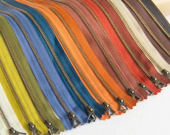 Könnte beinhalten: Eine Sammlung von 18 verschiedenen farbigen Reißverschlüssen mit Messing-Schiebern. Die Reißverschlüsse sind fächerförmig angeordnet, die Farben reichen von Beige bis Dunkelbraun.