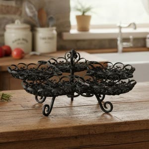 May include: Black, three-tiered decorative serving tray with ornate, scrolled metalwork. The tray has a central support and scrolled feet, ideal for displaying desserts or appetizers. The tray is on a wooden surface.