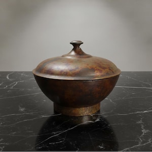 Op de afbeelding: Een bronzen kom met deksel, met een afgeronde body en een kleine, knopachtige handgreep aan de bovenkant. De kom staat op een korte, cirkelvormige basis. Het oppervlak heeft een gevlekt, verouderd uiterlijk, wat wijst op een vintage of antiek item. De kom staat op een zwart marmeren oppervlak.