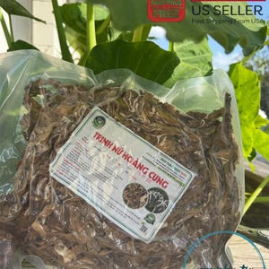 May include: A clear plastic bag filled with dried brown herbs labeled "TRINH NỮ HOÀNG CUNG" in Vietnamese. The bag is sitting on a wooden surface.