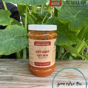 May include: A glass jar of red chili sauce with a white lid. The label is brown with white text that reads "Đặc sản Tây Ninh Muối Chu Hai Sốt Chấm Cay Vừa Dùng chay được Muối Chanh - Ăn là ghiền".