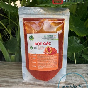 May include: A red pouch with a white label that reads "BỘT GẤC" and contains a red powder. The label also includes information about the product, such as the ingredients, usage, and storage instructions. The pouch is sitting on a green leafy background.