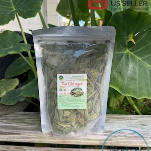 May include: A clear plastic bag filled with dried green leaves, likely herbs or tea. The bag has a label with Vietnamese text, including "Trà Cỏ ngọt." The background includes large green leaves and a wooden surface.
