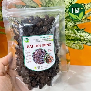 May include: A clear plastic bag filled with dark brown seeds. The bag has a green and white label with Vietnamese text and a logo. The label also includes a graphic of the seeds and leaves. The bag is held by a person.