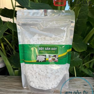 May include: A clear plastic bag filled with white tapioca starch granules. The bag has a green label with the text "BỘT SẮN ĐÂY 100% nguyên chất" in Vietnamese. The bag is sitting on a wooden surface.